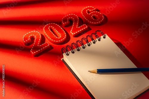 Red numbers 2026 are next to an open blank notebook and a blue pencil ready for writing down New year resolutions, future goals, and personal development plans on a vibrant red background