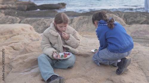 Two young girls are sitting on rocky seaside cliffs, enjoying a simple outdoor meal together. 