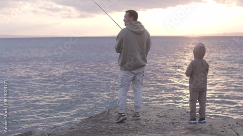 A father and son fish together by the sea at sunset, sharing a peaceful moment by the water. Dressed in hoodie
