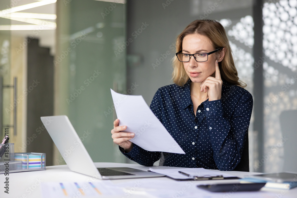 Fototapeta premium Young businesswoman wearing glasses focusing on financial documents while sitting at her desk with a laptop, clipboard, and pens in a contemporary office environment