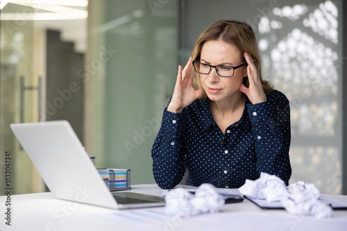 Businesswoman with glasses suffering from a headache and work related stress. Sitting at her desk with a laptop and crumpled papers. Feeling overwhelmed and exhausted in her modern office environment