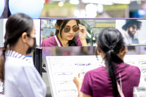 A young woman tries sunglasses at the mirror in an optical shop with the assistance of a saleswoman