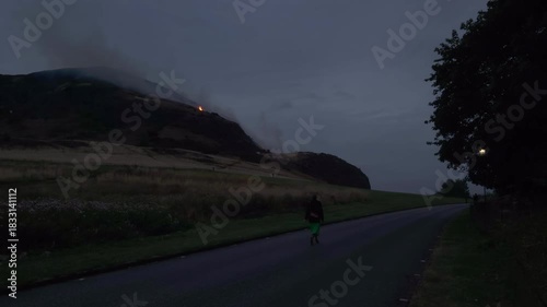 EDINBURGH, SCOTLAND, UK - AUGUST 10, 2025: A fire is visible on Arthur's Seat, Edinburgh, woman walks along a road nearby at dusk and then captures the blaze with a smartphone.