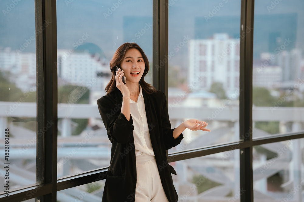 Fototapeta premium A professional woman standing by large windows, smiling while working on a laptop in a bright modern office environment.