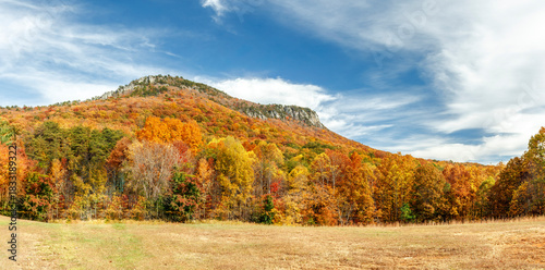 The steep, rocky cliffs of Cooks Wall, in Hanging Rock State Park in the North Carolina Sauratown Mountains, are surrounded by colorful fall foliage topped with a cloudy blue autumn sky.