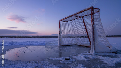Frozen ice hockey goal on a snowy lake at sunset with a hockey puck nearby