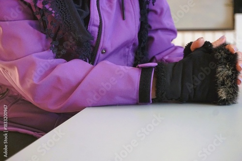 Schlanke Frau mit lila Winterjacke, schwarzem Schal und schwarzen Handschuhen sitzt auf Stuhl vor Tisch in Raum