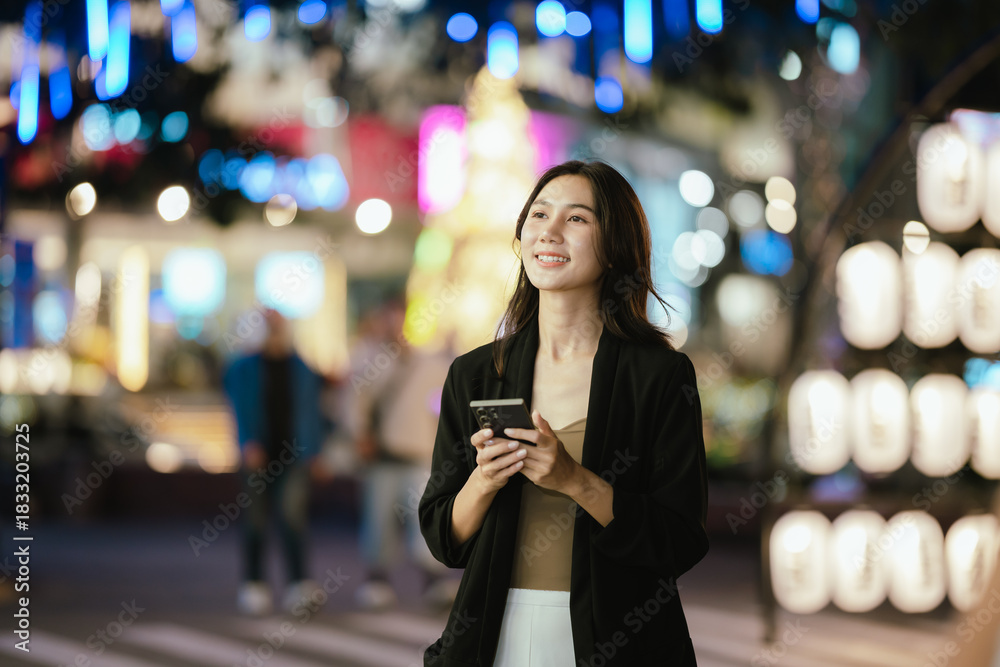 Fototapeta premium A smiling woman using her smartphone at night in a lively city setting, creating a warm and relaxed atmosphere with colorful blurred lights in the background.