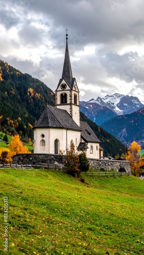 Alpine church atop green hill, amidst autumn leaves, with snow-capped mountains under a cloudy sky in the background