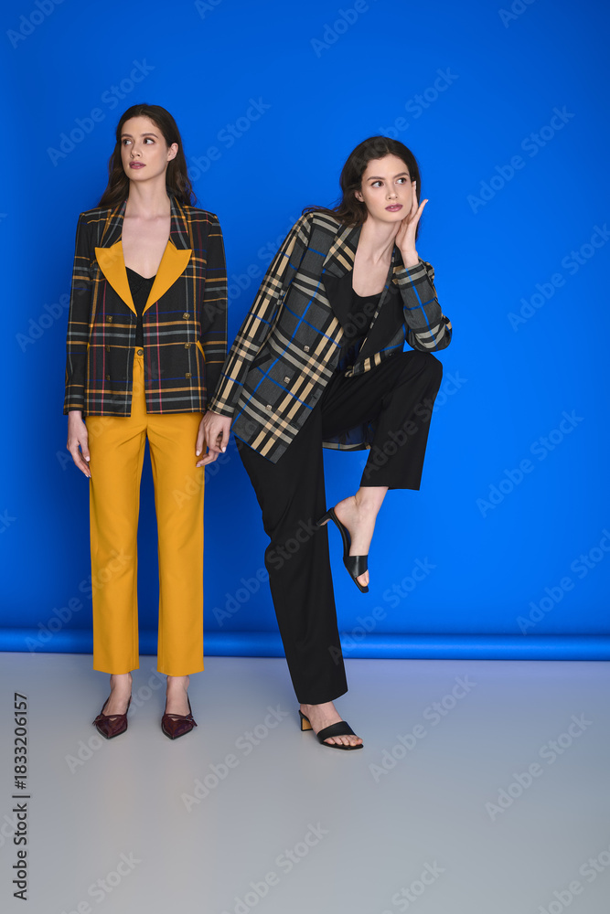 Fototapeta premium Young beautiful twin sisters showcase stylish attire against a vibrant blue backdrop