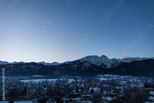 Fototapeta Naklejka Na Ścianę i Meble -  Snow-covered Zakopane nestled beneath the majestic, jagged, and dark peaks of the Tatra Mountains. A quiet, cold winter landscape at dawn, dusk