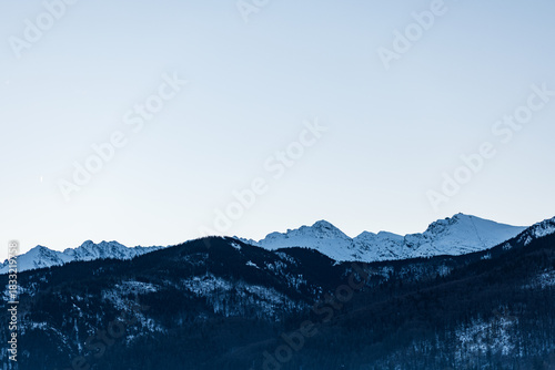 Fototapeta Naklejka Na Ścianę i Meble -  Serene winter mountain skyline with snow-covered peaks rising above dark, densely forested lower slopes under a pale, cold, gradient sky. Zakopane, Poland