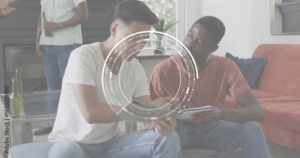 Fototapeta premium Collaborating men wearing white tee, rust tee leaning over tablet in living room, HUD, green bottle