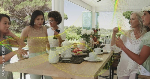 Gathering five adults wearing light green dress and white blouse at sunroom table, with teapots