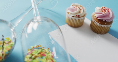 Showing two frosted cupcakes on card at pale blue surface, glasses spilling sprinkles, copy space
