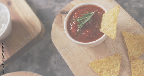 Showing white bowl of chunky tomato salsa with rosemary on wooden board, dark slate, nachos, salt
