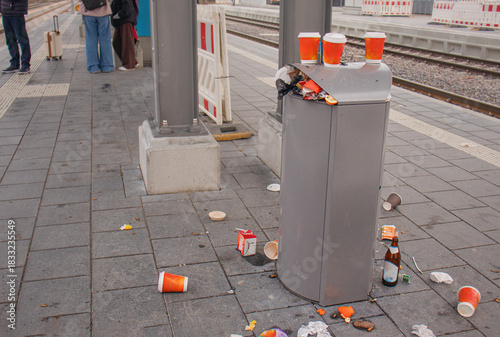 Overflowing trash bin with scattered litter on a train station platform, showing urban waste and pollution in a public area