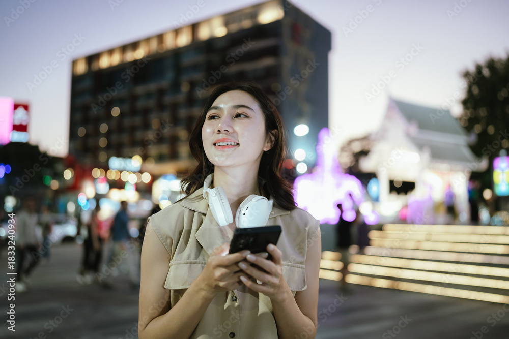 Fototapeta premium A young woman enjoying music with headphones while holding her smartphone, standing outdoors with colorful city lights glowing in the background.