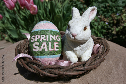 A decorative Easter display features a white bunny figurine and a painted egg with 'Spring Sale' text, both placed in a woven basket outdoors. Pink tulips and greenery appear in.