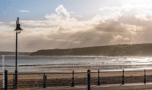 A promenade beside a beach with a headland in the distance and a cloudy sky.