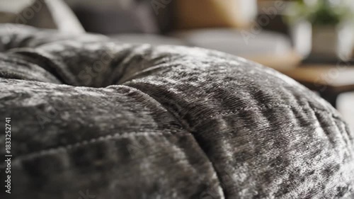 Close up view of a luxurious textured grey fabric ottoman with stitched details in a softly lit modern living room setting with a blurred background showing a sofa and coffee table