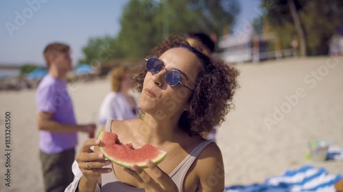 A young woman enjoying a slice of fresh watermelon on the beach while her friends relax and have fun in the background.