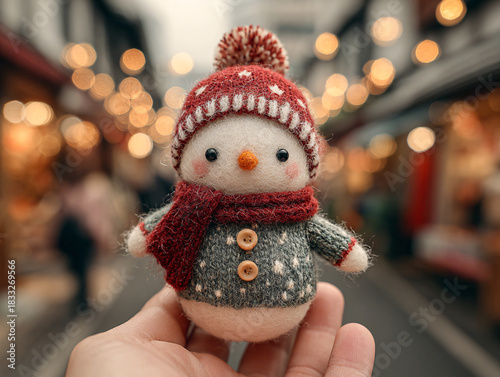 a small, knitted snowman toy held in someone's hand. The snowman is wearing a red and white striped hat, a red scarf, and red mittens. It has a carrot nose and is holding a small bundle of branches.