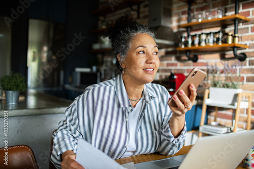 Mature woman smiling on speakerphone while working in home kitchen