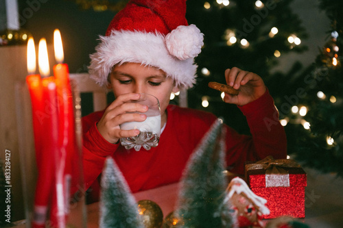 Funny toddler boy 7 y.o. in a Santa hat drinks milk and eats cookies in the kitchen with a decor for Christmas. Holidays, Traditions, baby food and health concept.