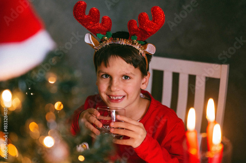 Toddler boy 7 y.o. in a red Santa hat drinks filtered water from a glass in the kitchen among the Christmas lights. Holidays, the concept of health.