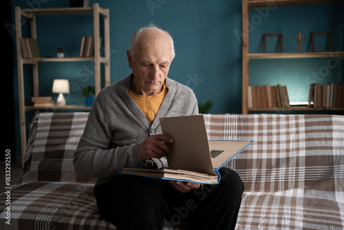 Lonely elderly widower sitting on couch looking at old photos recalling memories of deceased wife