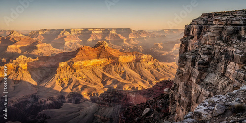 Panoramic view of vast, multi-layered canyon at sunset or sunrise, showing deep shadows and bright golden light on the rock formations.
