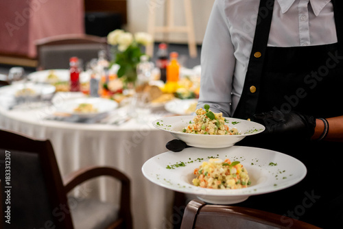 A waiter in a black apron and gloves serves two plates of a fresh, colorful vegetable salad in a restaurant setting with a white tablecloth table set in the background