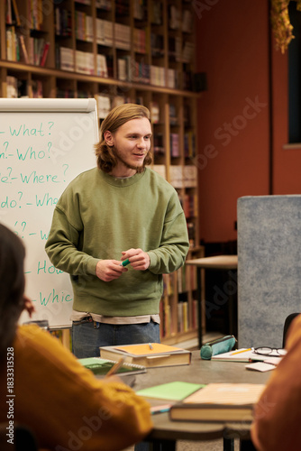 Young man standing in front of flip chart teaching group of people in classroom, holding marker and smiling, books and notebooks scattered on table in foreground
