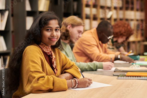 Portrait of teenage Asian girl smiling at camera while sitting at desk in library, multiethnic group of teenagers studying and writing in background, bookshelves