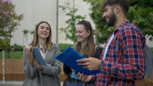 Three college students sit together on campus, holding books and having a friendly conversation. They enjoy the warm weather and talk casually while taking a break from studying.