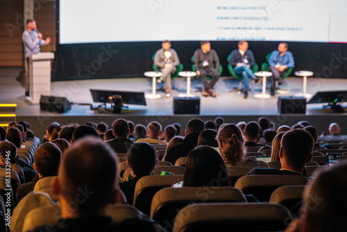 Panel of five speakers on stage during conference talk, audience listening intently in theater setting