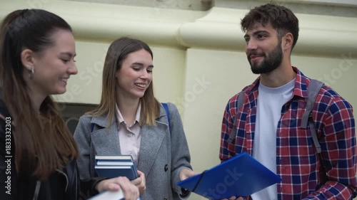 Three college students sit together on campus, holding books and having a friendly conversation. They enjoy the warm weather and talk casually while taking a break from studying.