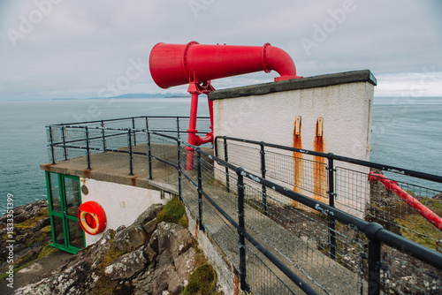 A striking detailed photograph of the iconic, bright red foghorn located at the base of the Ardnamurchan Lighthouse in the Scottish Highlands. The image highlights the vintage industrial design of the