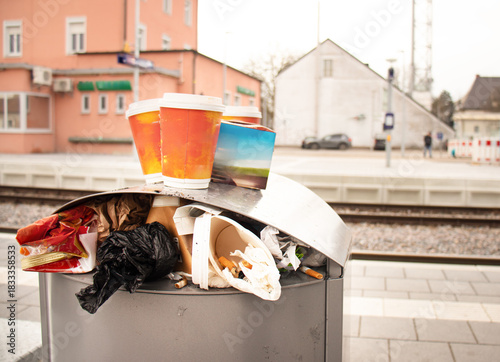 Overflowing trash bin with discarded coffee cups and litter on a train station platform, highlighting urban waste and pollution