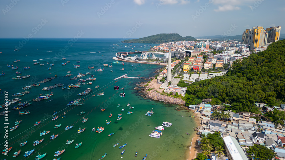 Fototapeta premium Aerial view of Phu Quoc's coastal town with colorful fishing boats, turquoise water, and the cable car in Vietnam. Scenic tropical seascape and vibrant waterfront in Vietnam.