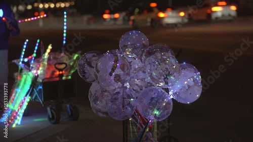 This video shows a close up of light up balloons and toys being sold by a street vendor at night, with traffic driving by in the background.