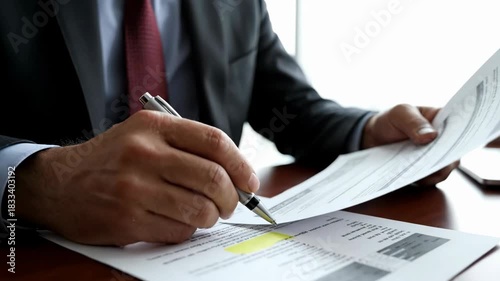 Man examining documents closely while highlighting important data. Bright office room with large windows and minimalistic design. Concept of corporate, finance, business consulting
