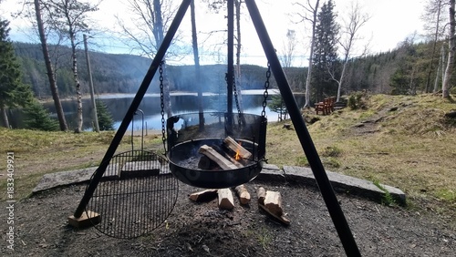 Scenic outdoor cooking setup by a peaceful lake in the forest with burning firewood during daytime
