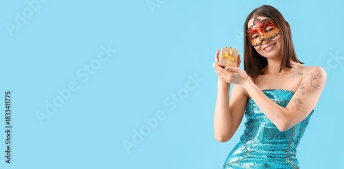 Beautiful young woman in carnival mask holding gift on blue background with s...