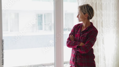 Young woman in red plaid pajamas opens the window, letting in fresh air. Her posture reflects relief and renewed energy. Concept of recovery, wellness, and fresh start. Back view, natural light, mid