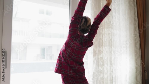 Young woman in red plaid pajamas opens the window, letting in fresh air. Her posture reflects relief and renewed energy. Concept of recovery, wellness, and fresh start. Back view, natural light, mid