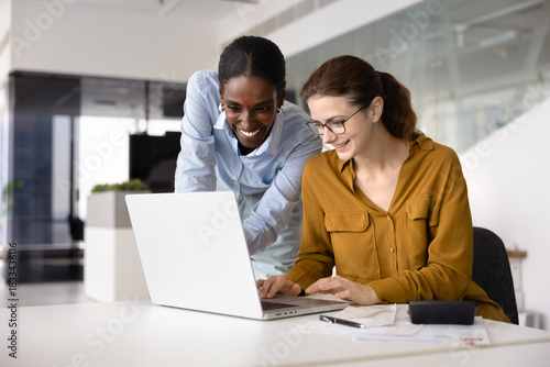 Two positive young diverse female colleagues working at laptop together