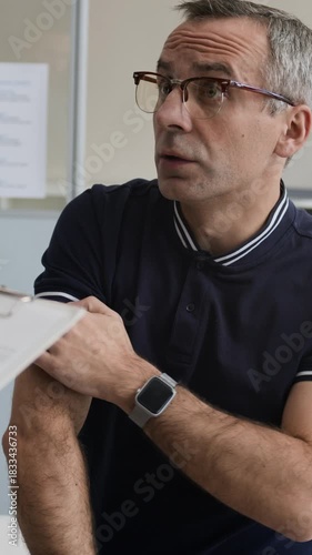 Vertical shot of troubled middle-aged Caucasian man with skin issues describing symptoms to unrecognizable doctor taking notes in outpatient card during appointment in dermatology clinic