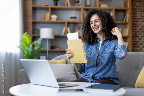 Happy young woman celebrating exciting news, opening an acceptance letter with a fist pump, sitting on a sofa with a laptop at home or in a student dormitory